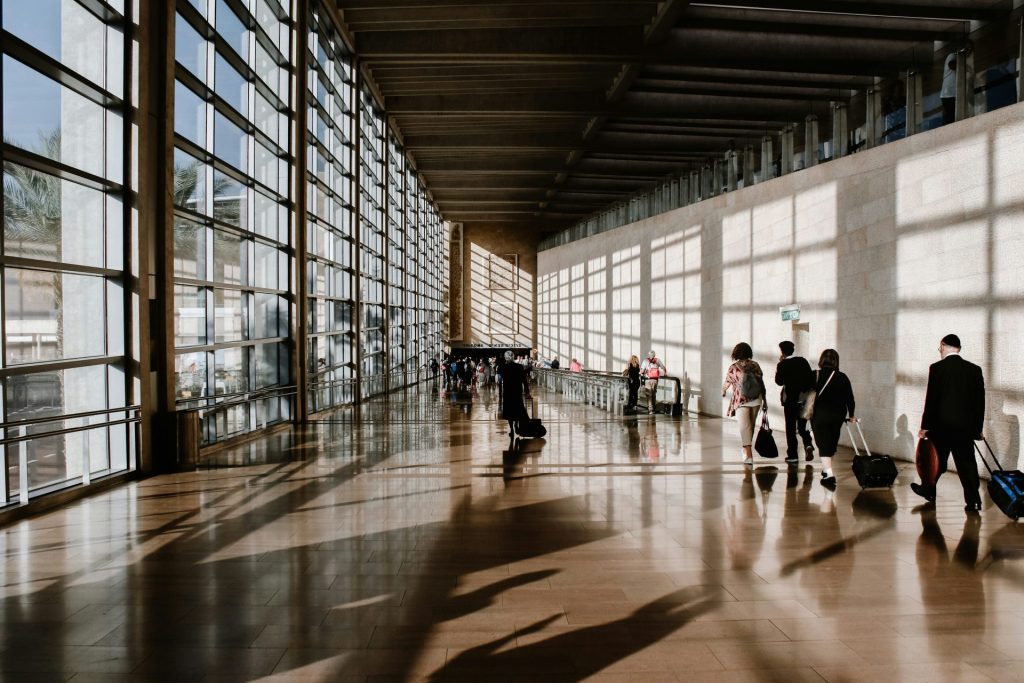 People walking through a sunlit airport terminal with large glass windows and shadows